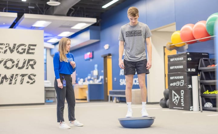 athletic-trainer-coaches-teen-athlete-on-balance-trainer-in-sports-facility