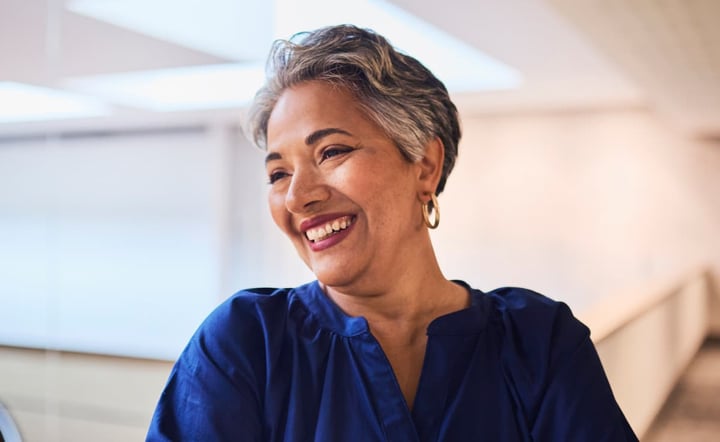 Close-up of a smiling mature woman with short grey hair and a blue blouse, arms crossed, in a bright, modern interior. 