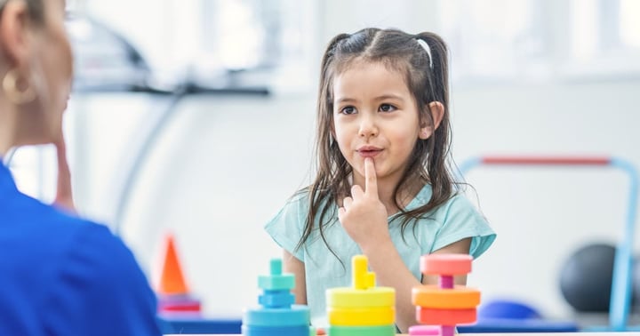 A female therapist sits with a sweet little girl as a table as they work on her speech together.