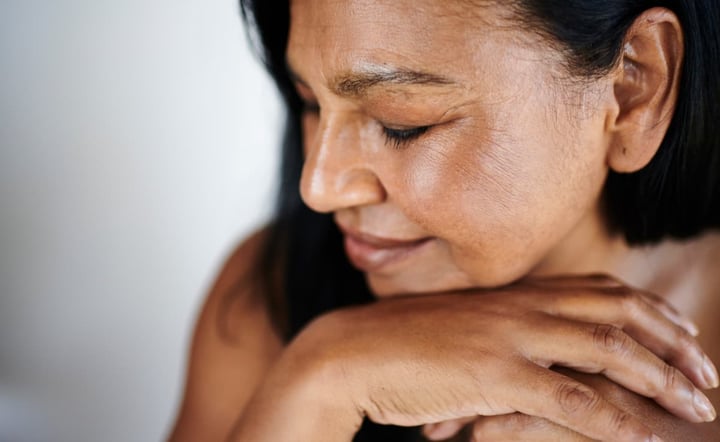 A serene mature woman of South Asian ethnicity sits with her eyes closed, hands gently resting on her chin, radiating calm and contentment in soft natural light.
