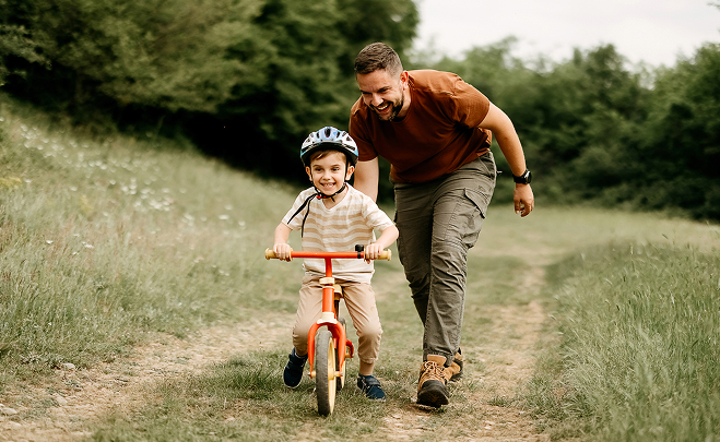 Father pushing child on bike on dirt and grass path