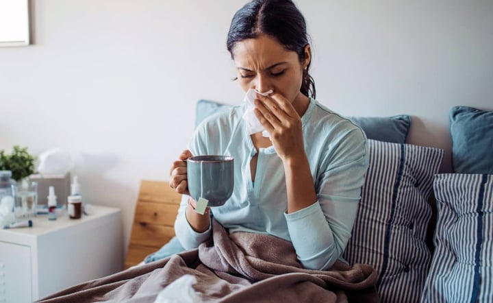 sick-woman-blowing-nose-with-tissue-while-drinking-tea-in-bed